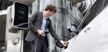 Below view closeup image of progressive black suit businessman recharge battery of his electric vehicle from public charging station. Renewable and alternative energy powered car concept.
