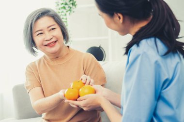 A young caregiver handing oranges to her contented senior patient at the living room. Senior care services, home visit by medical.