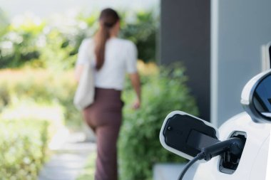 Focus EV charger plugged into EV car at home charging station with blurred background of progressive woman walking in background. Elective vehicle powered by clean energy for eco-friendly concept.