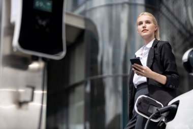 Businesswoman wearing black suit using smartphone, leaning on electric car recharge battery at charging station in city residential building with condos and apartment. Progressive lifestyle concept.