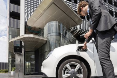 Below view closeup image of progressive black suit businessman recharge battery of his electric vehicle from public charging station. Renewable and alternative energy powered car concept.