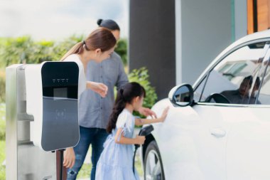 Progressive young parents and daughter with electric vehicle and home charging station. Green and clean energy from electric vehicles for healthy environment. Eco power from renewable source at home.