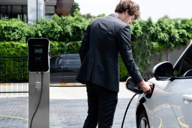 Modern şehir yerleşim bölgesindeki kamu şarj istasyonunda elektrikli araba şarj eden yenilikçi bir işadamı. Çevre dostu şarj edilebilir araba alternatif temiz enerjiyle çalışıyor..