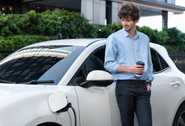 Progressive eco-friendly concept of parking EV car at public electric-powered charging station in city with blur background of businessman leaning on recharging-electric vehicle with coffee.
