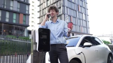 Takım elbiseli, hırslı bir işadamı elektrik kablosu ve yenilenebilir enerji enerjili bir elektrikli araçla şarj istasyonunda dururken elektrikli aracına yaslanıyor..