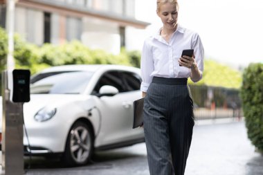 İş kadını tablet kullanıyor, halka açık otoparkta elektrikli aracını şarj ederken yürüyor. Teknolojinin ilerici yaşam tarzı ve EV otomobilinin ekolojik kaygısı