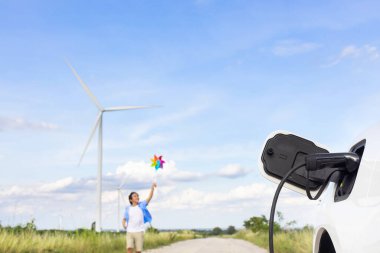 Progressive young asian boy playing with wind pinwheel toy in the wind turbine farm, green field over the hill. Green energy from renewable electric wind generator. Windmill in the countryside concept