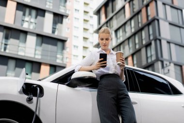 Focus businessman using phone, leaning on electric vehicle, holding coffee with blurred city residential condo buildings in background as progressive lifestyle by renewable and sustainable EV car.