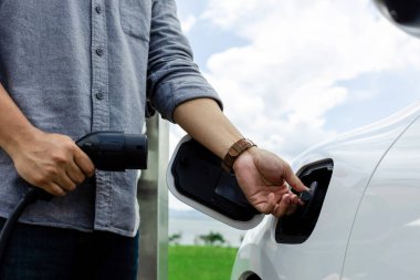 Progressive natural scenic where hand insert charging plug to electric vehicle from charging station with natural background. Future energy concept of EV car powered by sustainable electric energy.