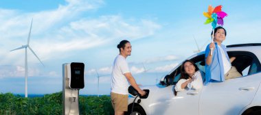 Concept of progressive happy family holding windmill toy and relax at wind farm with electric vehicle. Electric vehicle driven by clean renewable energy from wind turbine generator to charger station.