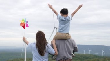 İlerici mutlu aile kavramı rüzgâr türbini çiftliğinde eğleniyor. Rüzgar türbini jeneratörüyle gelen elektrik jeneratörü kırsal bölgede tepe ve ufukta dağ var..