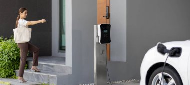 Asyalı bir kadın garajında uzaktan kumandalı araba anahtarıyla elektrikli arabayı kilitliyor, EV şarj istasyonunda şarj pili var. Alternatif temiz enerji günlük hayatta yenilikçi yaşam tarzı kavramı olarak uygulanır.