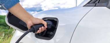 Progressive natural scenic with windmill generator where hand insert charging plug to electric vehicle from charging station with natural background. EV car powered by wind turbine electric generator.