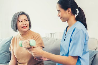 Caregiver helping contented senior woman exercise with dumbbell at home. Rehab for elder, senior medical care.