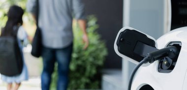 Arka planda bulanık baba ve kızının yürüdüğü şarj istasyonunda elektrikli araba şarj etmeye odaklan. Sağlıklı çevre yaşam tarzı konsepti için yenilikçi yeşil ve temiz enerji aracı.