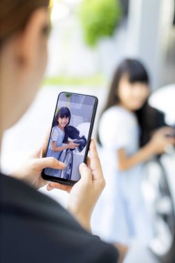 Akıllı telefon kullanan ilerici anne, EV 'yi şarj eden kızın odaklı fotoğrafını çekiyor. Asyalı bir kızın görüntüsü bulanık arkaplanlı telefon ekranında ev şarj istasyonundan EV vagonuna elektrikli şarj cihazı takıyor.