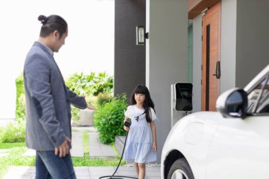 Progressive father and daughter plugs EV charger from home charging station to electric vehicle. Future eco-friendly car with EV cars powered by renewable source of clean energy.