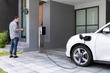 Progressive asian man and electric car with home charging station. Concept of the use of electric vehicles in a progressive lifestyle contributes to a clean and healthy environment.