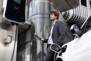 Below view of progressive businessman with electric car recharging at public charging station at modern city residential building. Eco friendly rechargeable EV car powered by alternative clean energy.