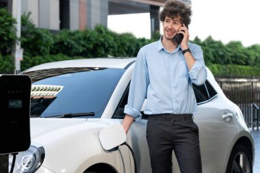 Progressive businessman talking on the phone, leaning on electric car recharging with public EV charging station, apartment condo residential building on the background as green city lifestyle.