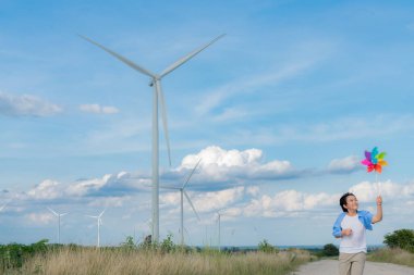 Progressive young asian boy playing with wind pinwheel toy in the wind turbine farm, green field over the hill. Green energy from renewable electric wind generator. Windmill in the countryside concept