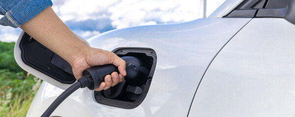 Progressive natural scenic with windmill generator where hand insert charging plug to electric vehicle from charging station with natural background. EV car powered by wind turbine electric generator.