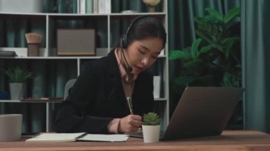 Kulaklık ve mikrofon takan iş kadını masasında çalışıyor ve dizüstü bilgisayar kullanıyor. Uzaktan kumandalı online toplantı için dizüstü bilgisayar kullanan hevesli kadın çalışanlar, ofiste iş videosu konferansı.