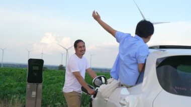 İlerici mutlu aile kavramı elektrikli araçla rüzgar çiftliğinde eğleniyor. Rüzgar türbini jeneratöründen gelen temiz yenilenebilir enerji ile şarj edilen elektrikli araç..
