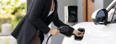 İlerici kadın elektrikli arabasının fişini şarj istasyonuna takıyor. İlerici bir yaşam tarzında elektrikli araç kullanımı kavramı temiz çevreye katkıda bulunur.