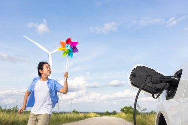 Progressive young asian boy playing with wind pinwheel toy in the wind turbine farm, green field over the hill. Green energy from renewable electric wind generator. Windmill in the countryside concept