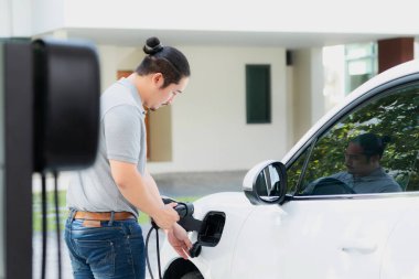 Progressive asian man install cable plug to his electric car with home charging station in the backyard. Concept use of electric vehicles in a progressive lifestyle contributes to clean environment.