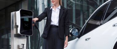 Progressive businesswoman wearing suit with electric car recharging at public parking car charging station at modern city center. Eco friendly rechargeable car powered by alternative clean energy.