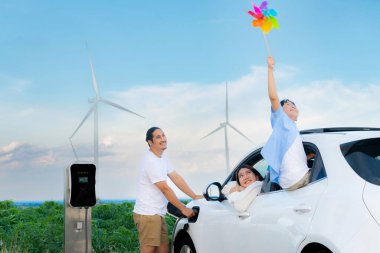 Concept of progressive happy family holding windmill toy and relax at wind farm with electric vehicle. Electric vehicle driven by clean renewable energy from wind turbine generator to charger station.