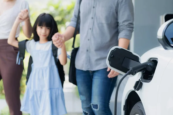 stock image Focus closeup electric vehicle recharging battery from home electric charging station with blurred family in background. Renewable clean energy car for progressive eco awareness lifestyle concept.