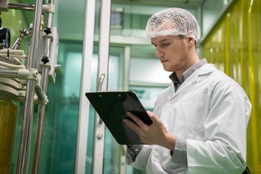 Eczacı bilim adamı bir CBD yağ çıkarıcısından bilgi almak için bir pano ve kalem kullanıyor ve tıbbi esrar ürünleri üretmek için bilimsel bir makine kullanıyor..