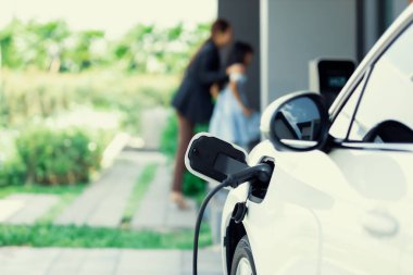 Focus EV car recharging at home charging station with blurred progressive woman and young girl in background for alternative clean energy technology concept for renewable electric vehicle.