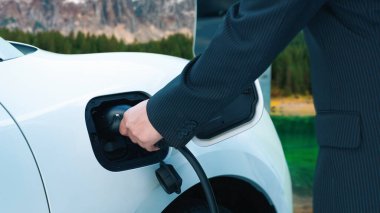 İlerici adam arka planda kırsal dağ olan şarj istasyonundan elektrik kablosu yerleştiriyor. Temiz ortam için elektro motor tarafından kullanılan EV otomobil