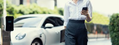 Kapalı alan iş kadını tablet kullanıyor, elektrikli aracını halka açık otoparkta şarj ederken yürüyor. Teknolojinin ilerici yaşam tarzı ve EV otomobilinin ekolojik kaygısı