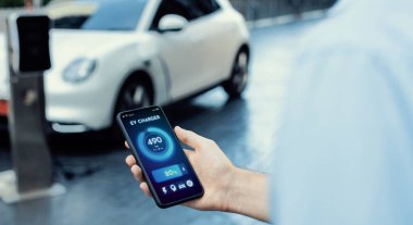 Closeup progressive businessman look at EV cars battery status application on smart phone screen at public parking charging station with power cable plug and renewable energy-powered electric vehicle