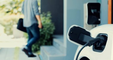 Focus electric car charging at home charging station with blurred progressive man walking in the background. Electric car using renewable clean for eco-friendly concept.
