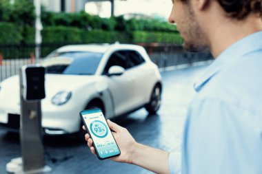 Closeup progressive businessman look at EV cars battery status application on smart phone screen at public parking charging station with power cable plug and renewable energy-powered electric vehicle