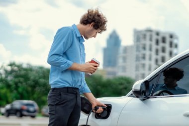 Progressive eco-friendly concept of parking EV car at public electric-powered charging station in city with blur background of businessman leaning on recharging-electric vehicle with coffee.