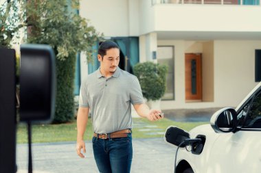 Progressive asian man install cable plug to his electric car with home charging station in the backyard. Concept use of electric vehicles in a progressive lifestyle contributes to clean environment.