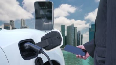 İlerici işadamı elektrikli aracı şarj kablosu ile şarj etme istasyonu ve temiz enerji kampanyası için şehir arka planına sahip akıllı telefondan enerji durumunu kontrol etme.