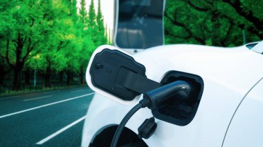 Çağdaş yeşil şehir ESG yeşil ağaçlar elektrikli araçla arka planda, EV araba şarj noktasındaki enerji yenilenebilir enerji taşımacılığıyla çevre bilincini artırmak için şarj ediliyor.