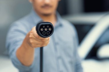 Focus on EV charging plug device from charging station for electric vehicle with blurred man holding plug and pointing at the camera background for alternative renewable energy.