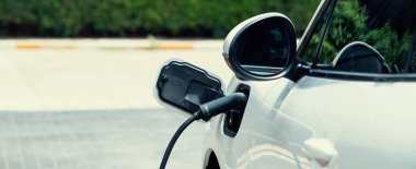 Closeup electric vehicle plugged in with EV charger at public charging station powered by renewable clean energy with residential building in background as progressive concept.