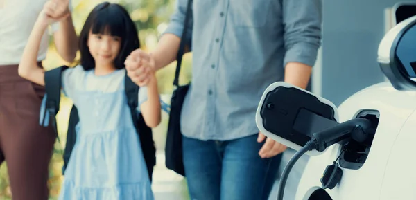 stock image Focus closeup electric vehicle recharging battery from home electric charging station with blurred family in background. Renewable clean energy car for progressive eco awareness lifestyle concept.