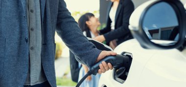 Closeup focus hand insert EV charger plug to electric vehicle at home charging station with blurred family in background. Progressive concept of green and clean energy to reduce CO2 emission by EV car