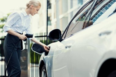 Progressive businesswoman insert charger plug from charging station to her electric vehicle with apartment condo building in background. Eco friendly rechargeable car powered by sustainable energy.